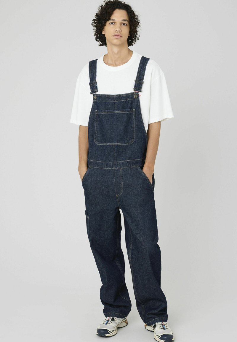 Salopette en jean bleu foncé avec bretelles réglables, poche avant et poches latérales. Associée à un t-shirt blanc et des chaussures de sport.