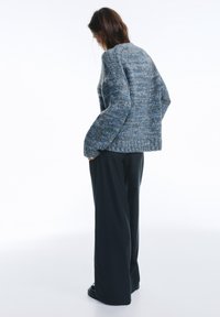 Grober, blauer Strickpullover mit strukturiertem Muster, weiten Ärmeln und lockerem Schnitt, kombiniert mit dunklen, Nadelstreifen, weiten Hosen.