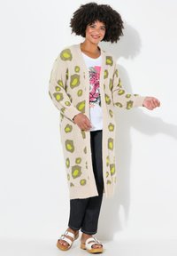 Beige cardigan dengan pola leopard hijau dan coklat, bagian depan terbuka, lengan panjang, tekstur rajutan, dipasangkan dengan kaos grafis dan jeans gelap.