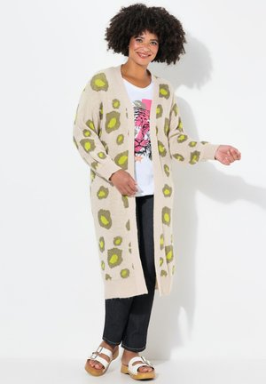 Gilet beige avec un imprimé léopard vert et marron, devant ouvert, manches longues, texture tricotée, associé à un t-shirt graphique et un jean foncé.