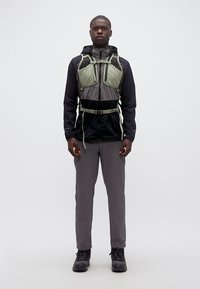 Άνδρας που στέκεται φορώντας μαύρο και γκρι εξωτερικό μπουφάν με πράσινο θώρακα και γκρι παντελόνι, κοιτάζοντας προς τα εμπρός σε λευκό φόντο.