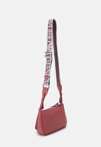 Červená krosna z texturované kůže s odnímatelným popruhem zdobeným logem a pruhy. Zapínání na zip, obdélníkový tvar a přední kapsa.