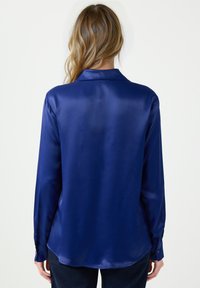 Granatowa jedwabna koszula o gładkiej fakturze, z długimi rękawami i kołnierzykiem. Widok z tyłu pokazuje zaokrąglony dół i mankiety zapinane na guziki.