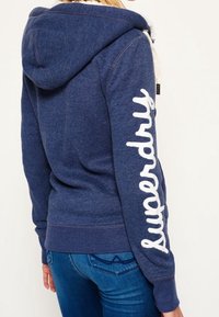 Felpa blu navy con una texture morbida, caratterizzata da un logo bianco "Superdry" sulla manica sinistra e un cappuccio con cordino. Stile fitted.