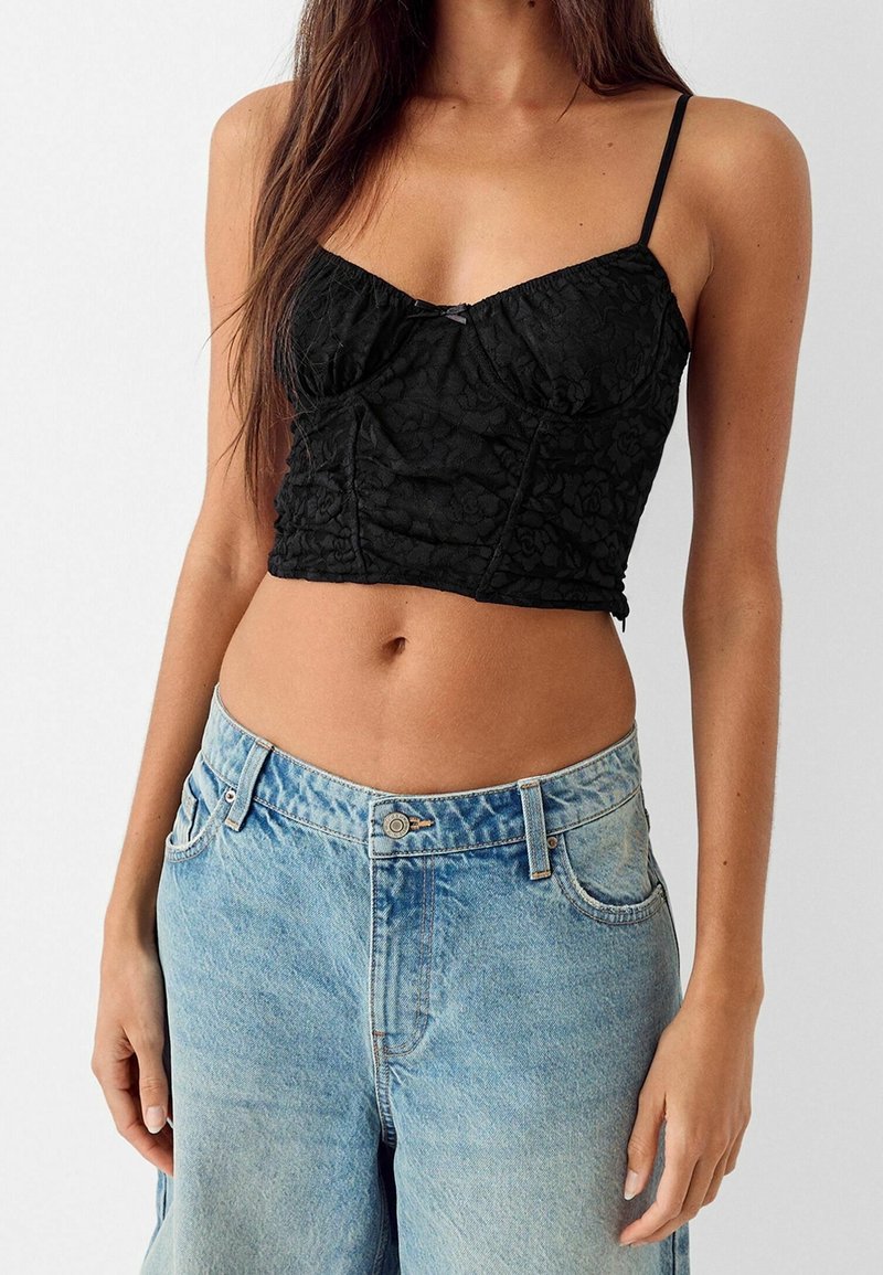 Musta pitsinen lyhyt toppi, jossa on säädettävät spagettiolkaimet, kukkakuviot ja rypytysdetaljit, yhdistettynä vaaleansinisiin löysiin farkkuhousuihin.