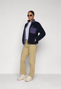 Lunt fleecejakke i marineblå med en lilla lomme, beige bukser og lyse sneakers. Jakken har høy krage og glidelås.