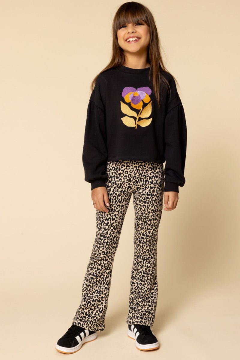 Zwarte cropped sweatshirt met een paarse en gouden bloemenontwerp, gecombineerd met leopardenprint flared broeken en zwarte sneakers met witte strepen.