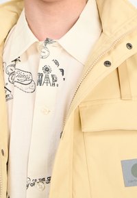 Giacca gialla chiara con zip, dotata di tasca laterale e etichetta. Sotto c'è una camicia crema con stampe grafiche nere e bottoni.