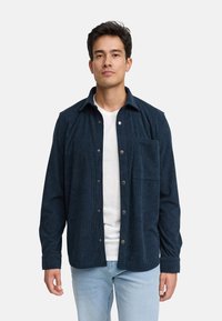 Mørkeblå kordfløyelskjorte med knappelukking foran, enkelt brystlomme og lange ermer, lagd over en hvit t-skjorte og kombinert med lyse jeans.