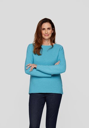 Helles, strukturiertes blaues Sweatshirt mit langen Ärmeln, Rundhalsausschnitt und lockerer Passform. Kombiniert mit dunklen Jeans, vor einem grauen Hintergrund stehend.