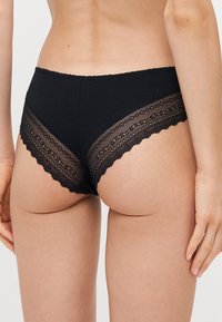 Schwarze Baumwollunterwäsche mit Spitzenverzierungen an den Seiten, die einen gewellten Rand und geometrische Muster aufweisen. Weicher Stoff und mittlere Leibhöhe.