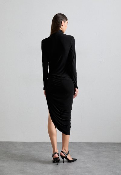 Rochie neagră cu mâneci lungi, cu detalii plisate, până la genunchi, cu guler înalt și slit pe o parte, asortată cu pantofi cu toc înalt negri.