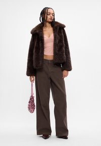 Casaco de pele sintética castanho com um colarinho largo, top justo rosa, calças largas castanhas e uma bolsa texturizada rosa. Texturas suaves e macias por todo o lado.