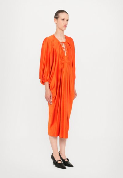 Vestido midi de satén naranja con mangas abullonadas y detalle de lazo en el escote. Combinado con zapatos de tacón negro puntiagudos. Textura suave y diseño drapeado.