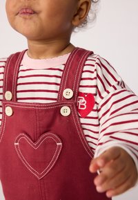 Kleinkind trägt rote Latzhose mit Herzstickerei und gestreiftem Langarmshirt, lehnt sich nach vorne mit leicht aufgereihten Lippen.