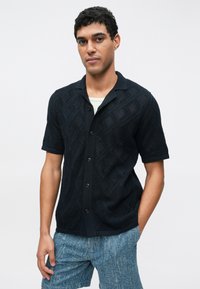 Zwart overhem met korte mouwen en knoopsluiting, voorzien van een textuur met ruitpatroon, een contrasterende kraag en een comfortabele pasvorm, gecombineerd met lichte denim shorts.