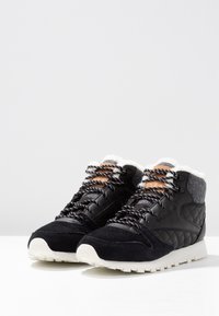 Ett par svarta vintersneakers med vita sulor, vadderade sidor, mönstrade snören och mjukt fleecefoder vid ankla kragen.