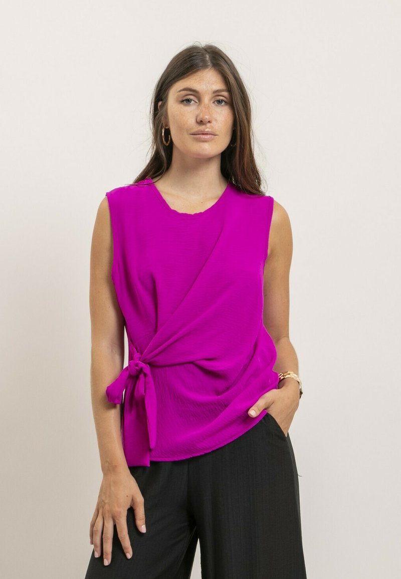 Top sans manches fuchsia avec un nœud noué à la taille, présentant une texture subtile et un col rond, associé à un pantalon noir.