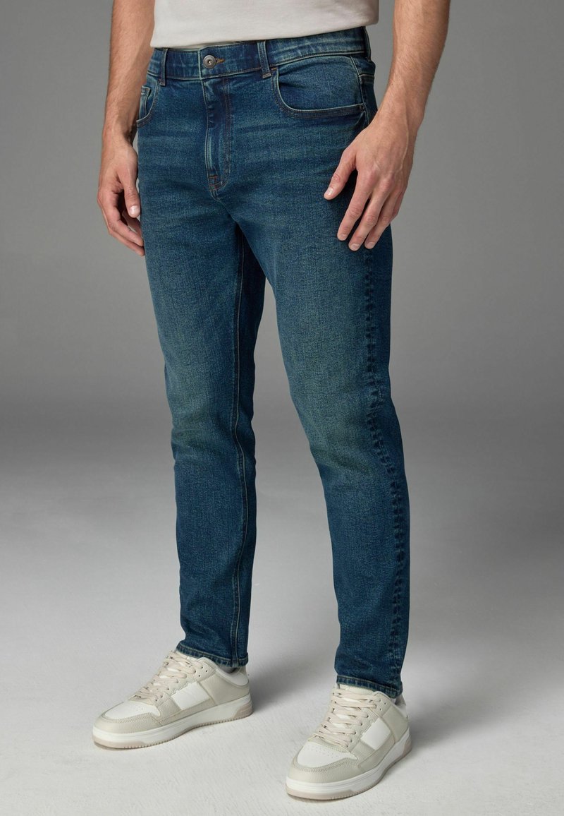Jeans de denim bleu cu o croială slim, dotate cu buzunare frontale, închidere cu buton și ușoare decolorări. Potrivite cu adidași albi de sport.