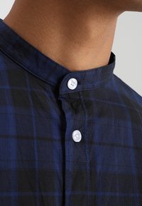 Camicia a quadri blu marina con colletto a fascetta, bottoni bianchi e tessuto di cotone leggero. La parte ravvicinata mostra i dettagli del colletto e dei bottoni.