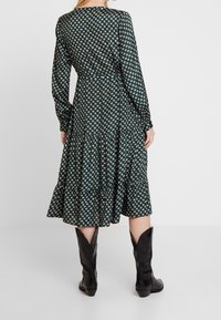 Groene jurk met patroon, lange mouwen, een gekruide zoom en een aansluitende taille. Draag met zwarte kniehoge laarzen. De stof lijkt glad en lichtgewicht.