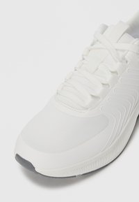 White athletic shoe with textured sole, mesh upper, and crisscross laces, shown from front and side angle on a plain background.