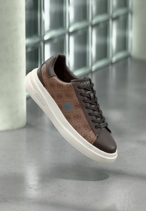 Brązowy i ciemnobrązowy niski sneaker z białą podeszwą, wzorzystymi bokami i logo "GUESS" na języku i boku, unoszący się nad szarą podłogą.