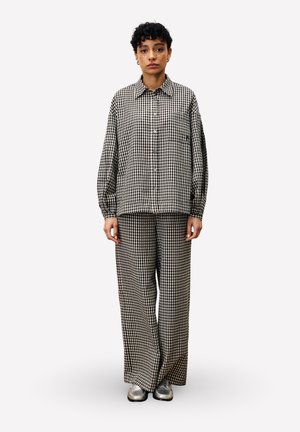 Femme debout portant une chemise ample à carreaux noir et blanc et un pantalon large assorti, avec des chaussures argentées, sur un fond uni.