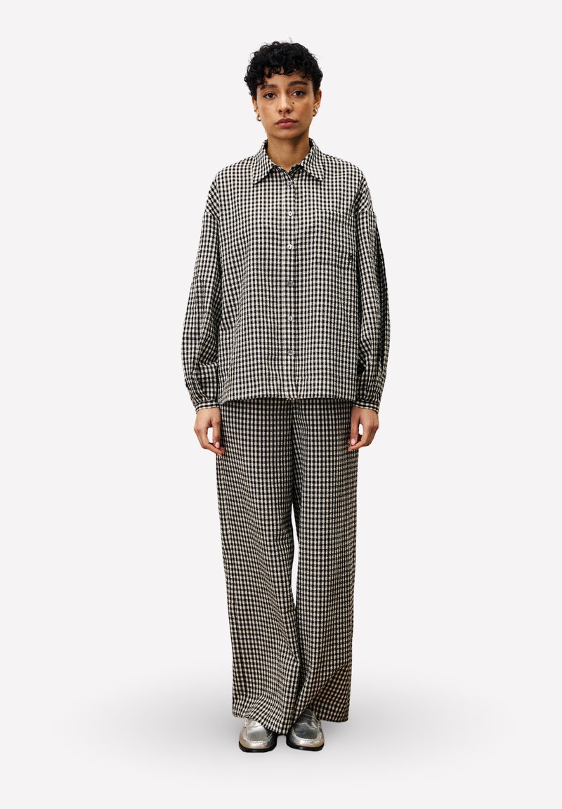Vrouw die staat in een zwart-wit geblokt los shirt en bijpassende wijde broek met zilveren schoenen tegen een effen achtergrond.