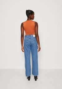 Rustfärgad ärmlös topp i kombination med ljusblå vida jeans. Jeansarna har en vit etikett på bakre midjan och två bakfickor.