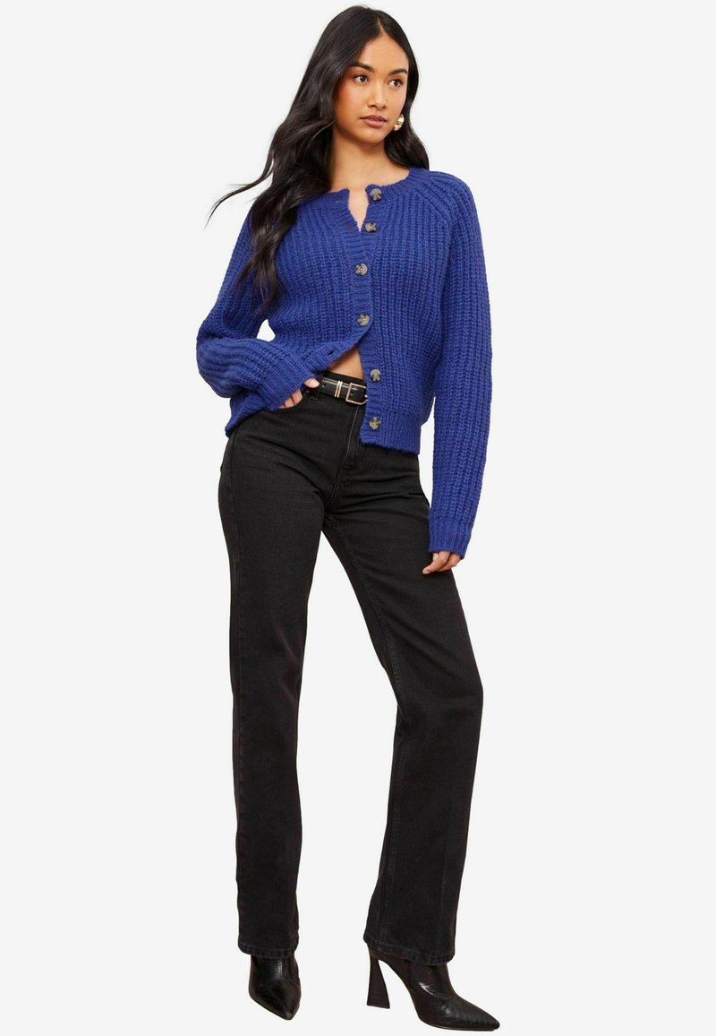 Mėlyna megzta kardiganas su tekstūruotu raštu, su šešiais mygtukais priekyje, derinama su juodomis tiesiomis kelnėmis ir aukštakulniais.