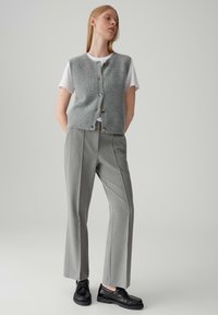 Gilet grigio a maglia con bottoni sopra una t-shirt bianca, abbinato a pantaloni grigi sartoriali e scarpe nere. Texture liscia e design aderente.