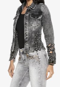 Musta ja harmaa farkkutakki, jossa on monimutkaiset pitsikoristeet ja koristelut hihoissa ja sivuilla, napillinen suljenta ja kaksi etutaskua.