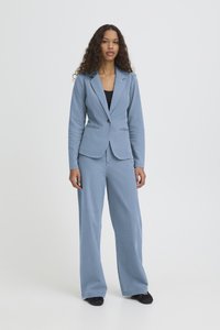 Femeie cu păr creț în picioare, purtând un blazer albastru deschis și pantaloni largi asortați peste un top negru, pe un fundal simplu deschis la culoare.