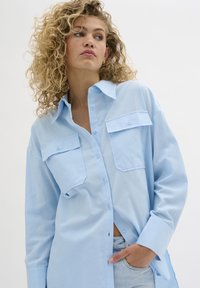 Light blue button-up shirt in soft fabric, featuring two chest pockets, long sleeves, and a relaxed fit with a slightly curved hem.