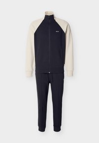 Φόρμα ναυτικού μπλε με κρεμ λεπτομέρειες· ζακέτα με φερμουάρ και ψηλό γιακά με λογότυπο, συνδυασμένη με στενά παντελόνια με κορδόνι και τσέπες με φερμουάρ.