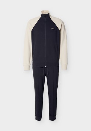 Survêtement marine avec accents crème ; veste zippée à col montant et logo, associée à un pantalon fuselé doté d'un cordon de serrage et de poches zippées.