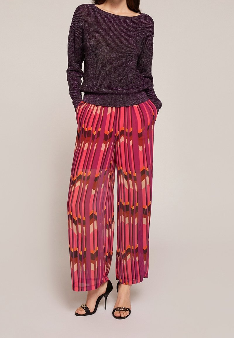 Femme portant un pull à manches longues de couleur violet foncé, un pantalon large à motifs multicolores roses et des sandales noires à talons hauts.