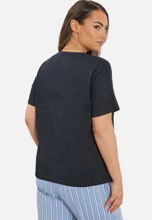 Kvinde iført en ensfarvet sort kortærmet skjorte og blå stribede bukser, kigger over sin skulder mod en hvid baggrund.