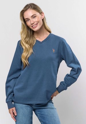 Blå V-hals sweatshirt lavet af blødt stof, med lange ærmer og en lille broderet logo på venstre bryst. Kombineret med lyse jeans.