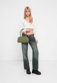 Mala de tecido verde com alças duplas, com detalhes em prata; modelo veste um suéter branco curto e jeans de corte largo.