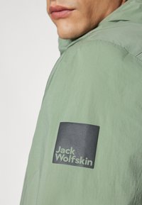 Lysegrøn jakke med en glat overflade, der har et sort firkantet logo-patch, som står "Jack Wolfskin" i hvid tekst på ærmet.