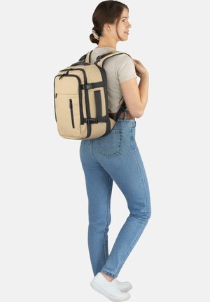 Beige nylon backpack with black accents, multiple compartments, padded straps, and a sturdy design, worn by a person in light jeans.