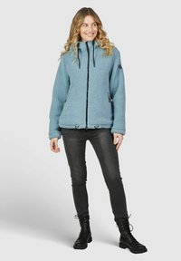 Veste en polaire bleu clair avec une fermeture éclair intégrale, un col haut et un cordon de serrage ajustable. Accents noirs sur les fermetures éclair et les poignets. Jean noir ajusté.