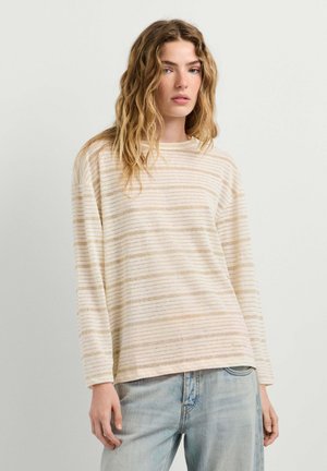 Junge Frau mit welligem Haar, die ein beige-weiß gestreiftes Langarmshirt und hellblaue Jeans vor einfachem Hintergrund trägt.