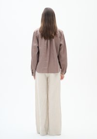 Bruine linnen blouse met lange mouwen en schouders die naar beneden vallen, gecombineerd met lichtbeige wijde linnen broek, gezien vanaf de achterkant tegen een witte achtergrond.
