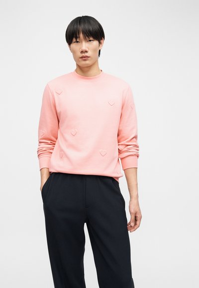 Νέος άνδρας που φοράει ροζ φούτερ με μικρά μοτίβα καρδιών, μαύρο παντελόνι, στέκεται με τα χέρια στις τσέπες μπροστά σε ένα απλό φόντο.