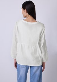Hvid, struktureret bluse med en afslappet pasform, rund hals og 3/4-ærmer. Båret sammen med lyseblå jeans, som viser et afslappet look fra ryggen.