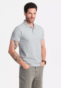Camisa polo gris claro de manga corta, con una tapeta de tres botones y un pequeño logo en el lado izquierdo del pecho, combinada con pantalones de color claro.