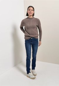 Brązowy sweter w warkocze połączony z ciemnoniebieskimi skinny jeansami i białymi, niskimi sneakersami z jasnoniebieskimi akcentami. Proste tło.